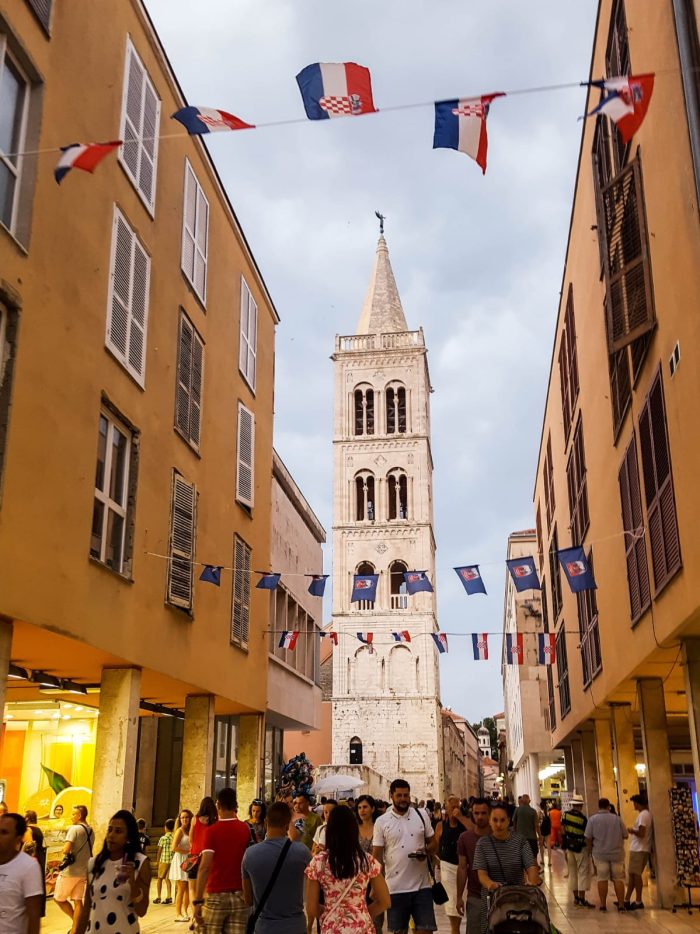 People walking along the historic streets of Zadar, Croatia, with local shops and the Adriatic Sea in the background