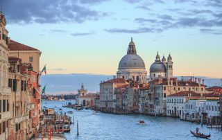 A panoramic luxury view of Venice’s winding canals with historic buildings and boats under a clear sky. contact us now